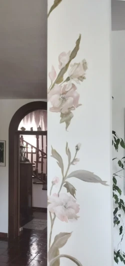 A modern living room with a striking floral mural painted on the main wall. The mural features large, detailed flowers in various shades of pink, red, and orange, with lush green leaves intertwined. The vibrant colors of the floral design contrast beautifully with the neutral tones of the room's furnishings. Below the mural, a sleek, white sofa with colorful throw pillows sits on a light wooden floor, creating a cozy seating area. A minimalist coffee table and a soft area rug in complementary colors complete the space. Natural light streams in through a nearby window, highlighting the artistic floral wall and enhancing the room's bright, inviting ambiance. Make your space unique and original with handpainted creative decorations on wall